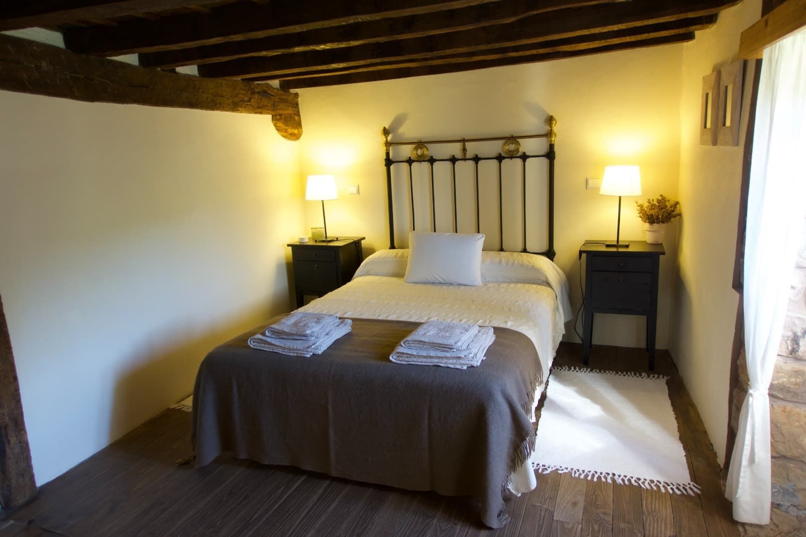 Dormitorio con cama, mesitas de noche, lámparas, vigas de madera y alfombra blanca.