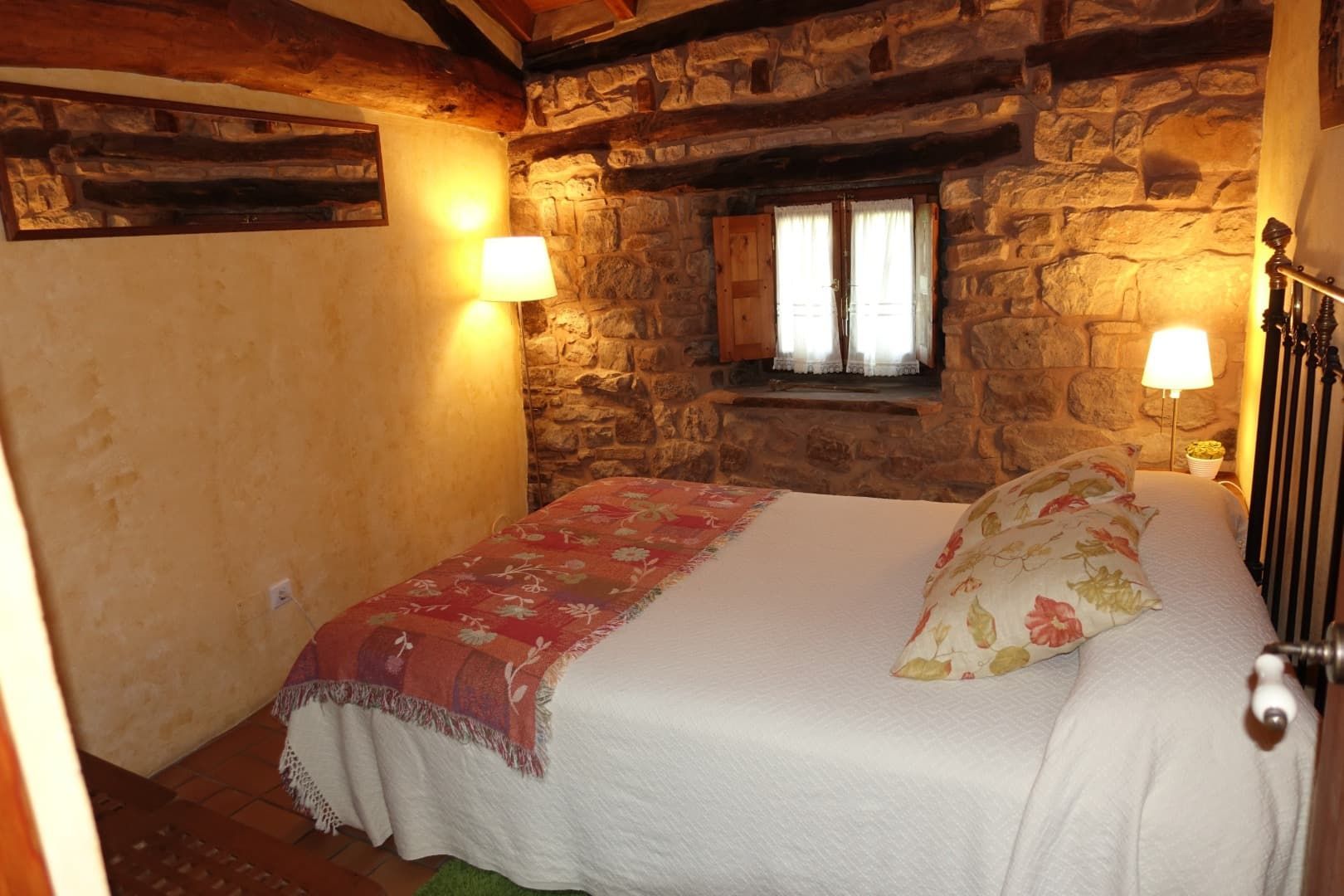 Dormitorio con paredes de piedra, ropa de cama blanca, manta floral, pequeña ventana y lámparas.