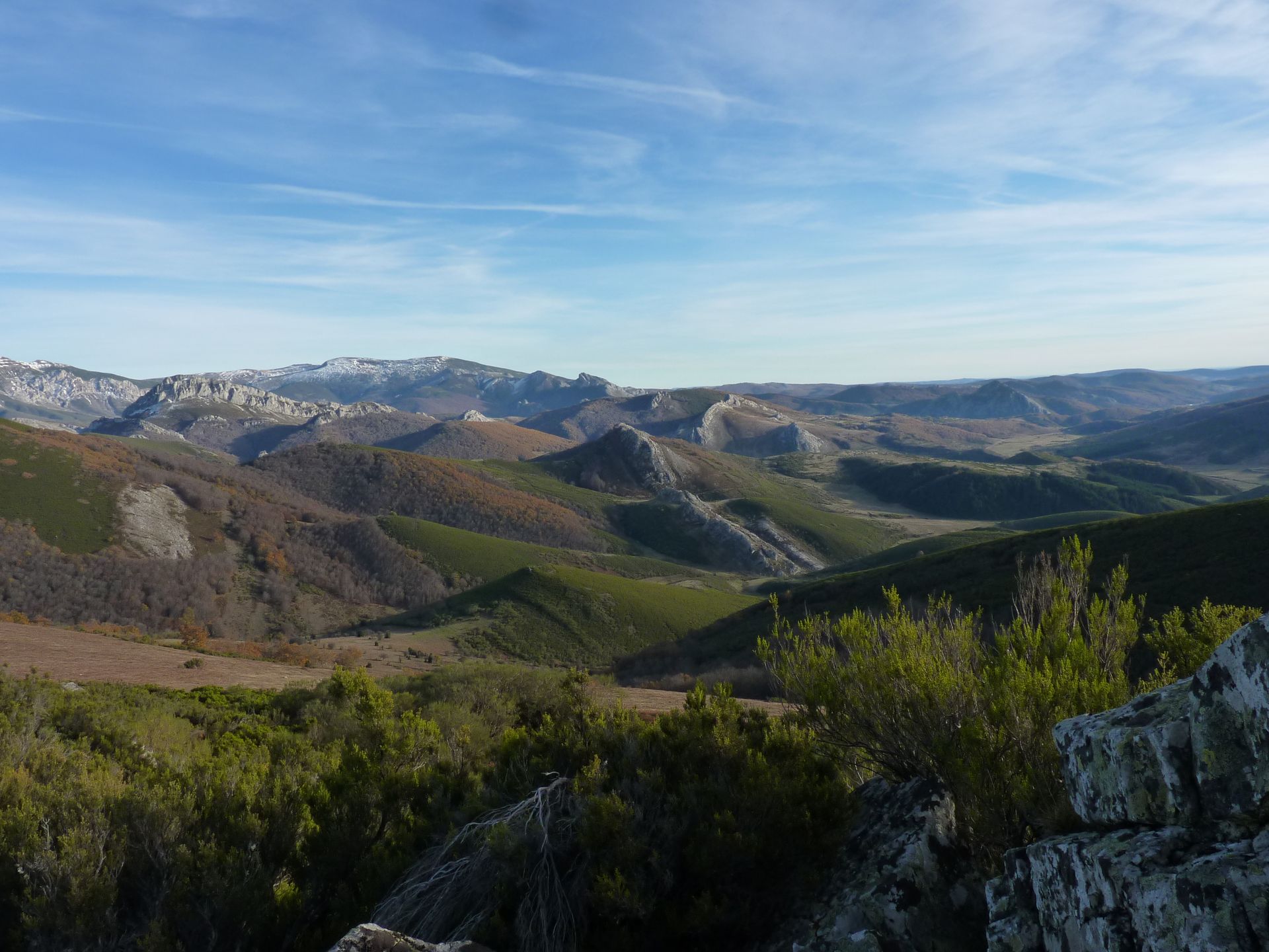 Paisaje montañoso ondulado con vegetación verde y marrón bajo un cielo azul.