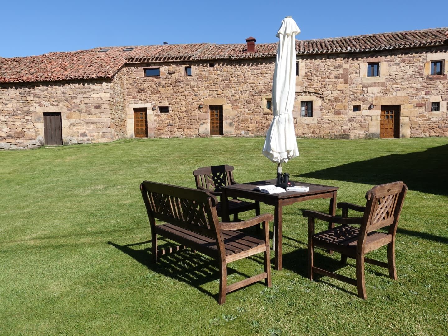 Muebles de patio exterior sobre césped frente a un edificio de piedra con puertas abiertas. Sombrilla blanca.