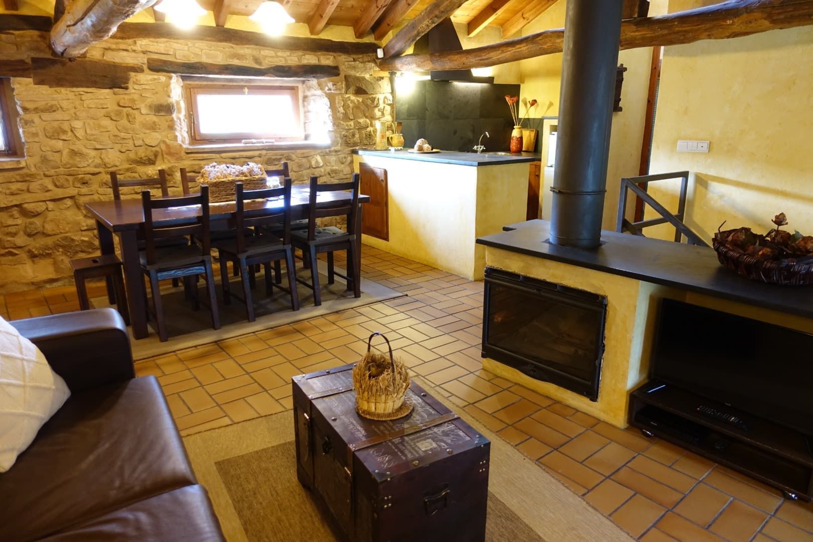 Espacio habitable rústico con paredes de piedra, vigas de madera, mesa de comedor, chimenea y cocina.
