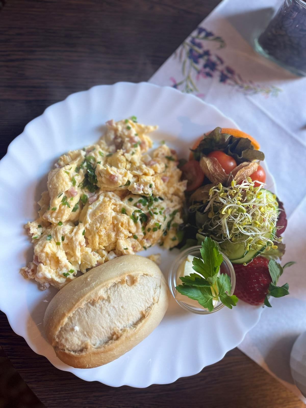 Ein weißer Teller mit Rührei, einem Brötchen und einem Salat.