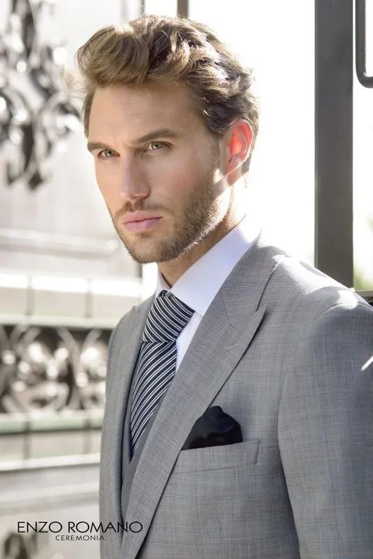 Hombre con traje gris, camisa blanca y corbata a rayas, posando al aire libre.