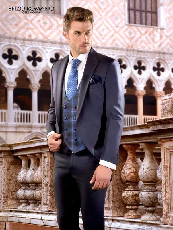 Hombre con un traje azul marino, chaleco a cuadros, corbata azul claro y pañuelo de bolsillo, de pie frente a un edificio.