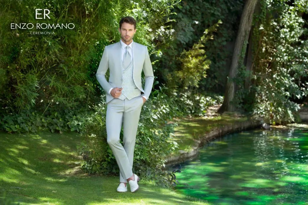 Un hombre con traje verde claro se encuentra cerca de un estanque con zapatillas blancas y vegetación verde en el fondo.