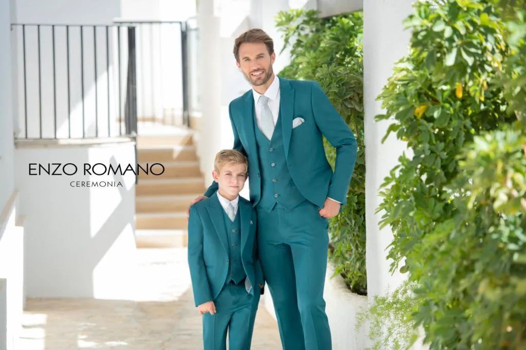 Hombre y niño con trajes verde azulado, sonriendo, de pie afuera de un edificio con escaleras y follaje.