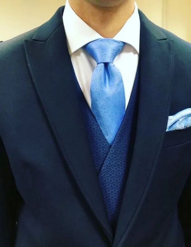 Hombre con traje azul marino, corbata, chaleco y pañuelo de bolsillo azul claro. Camisa blanca.