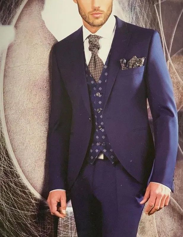Hombre con traje azul, chaleco estampado y corbata, apoyado sobre un fondo neutro.