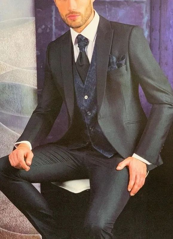 Hombre con traje oscuro, chaleco y corbata, sentado. Camisa blanca y pañuelo de bolsillo. Fondo morado.