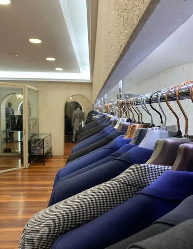 Hilera de chaquetas de traje en varios tonos de azul colgadas en una tienda de ropa bien iluminada. Suelo de madera.