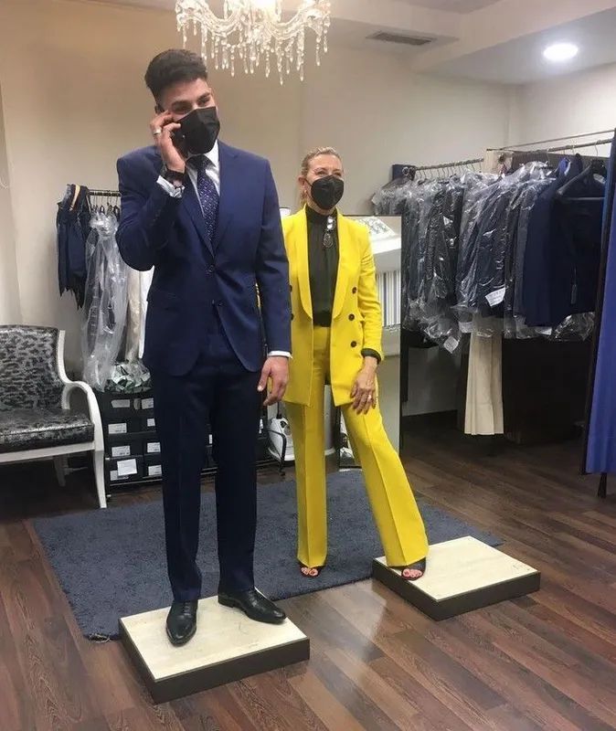 Dos personas vestidas formalmente y con máscaras, paradas sobre plataformas en una tienda de ropa.