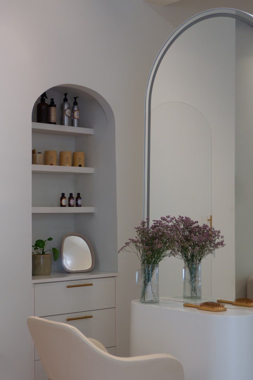 Un espacio de peluquería minimalista con una estantería arqueada empotrada, productos, un tocador, un espejo grande y un jarrón con flores.