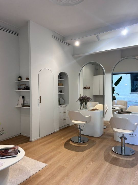 Un salón luminoso y minimalista con espejos blancos en forma de arco, sillas blancas modernas y suelo de madera clara.