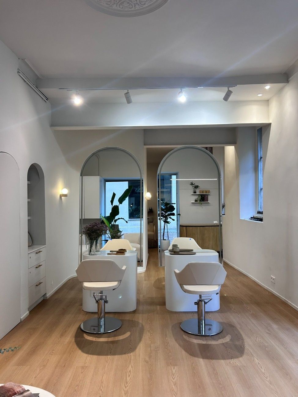 Interior de peluquería con dos sillas frente a espejos, puertas arqueadas, paredes blancas y piso de madera.