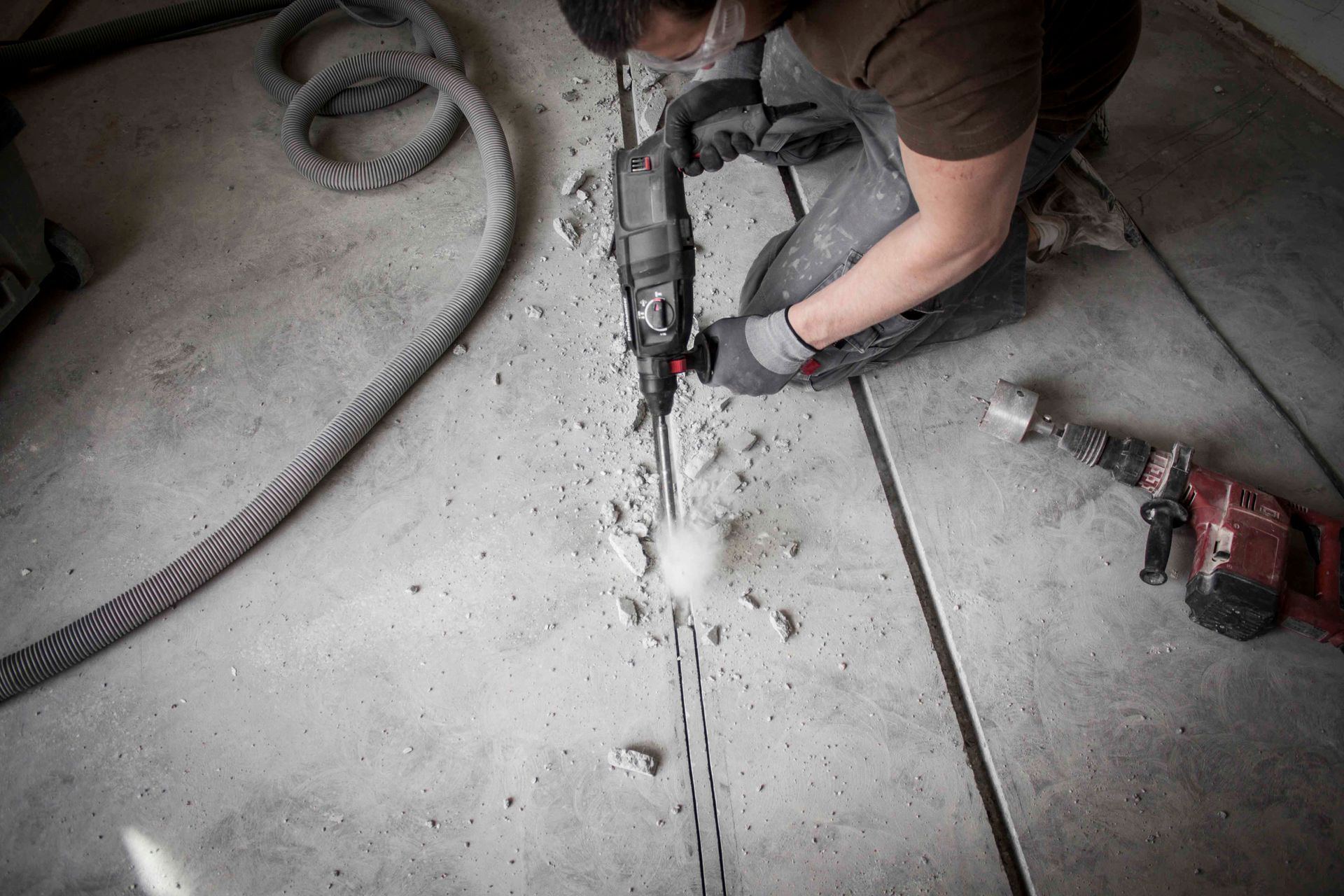 Ein Arbeiter schneidet mit einem Elektrowerkzeug in einen Betonboden; in der Nähe befinden sich Staub und eine Bohrmaschine.