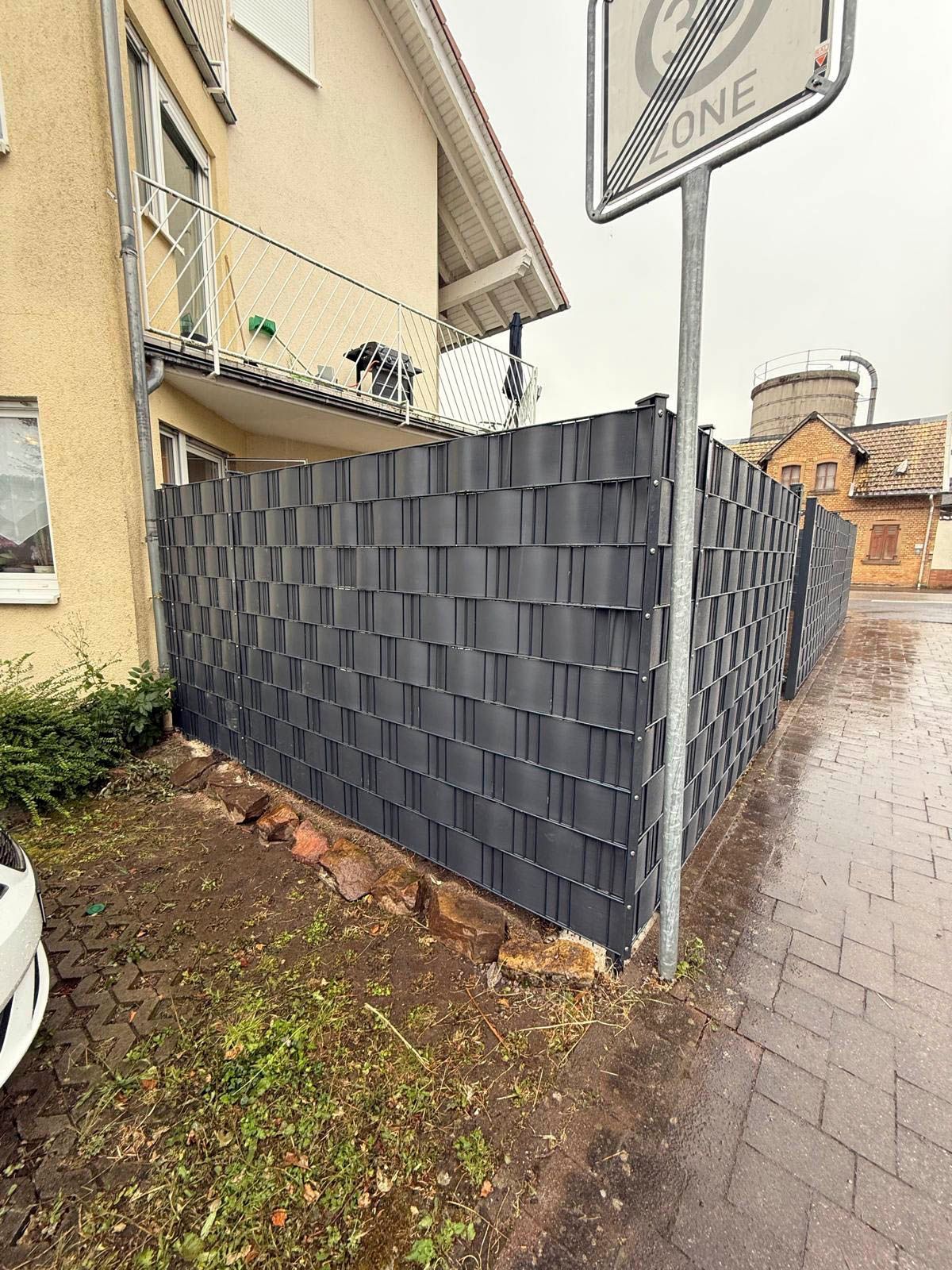 Dunkelgraues, gewebtes Zaunelement neben einem beigen Haus auf einem nassen Wohngebietsbürgersteig