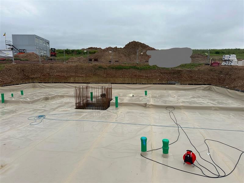 Betonbaustelle mit Bewehrungssäule, grünen Rohren und roten Geräten auf einer flachen Betonplatte unter bedecktem Himmel