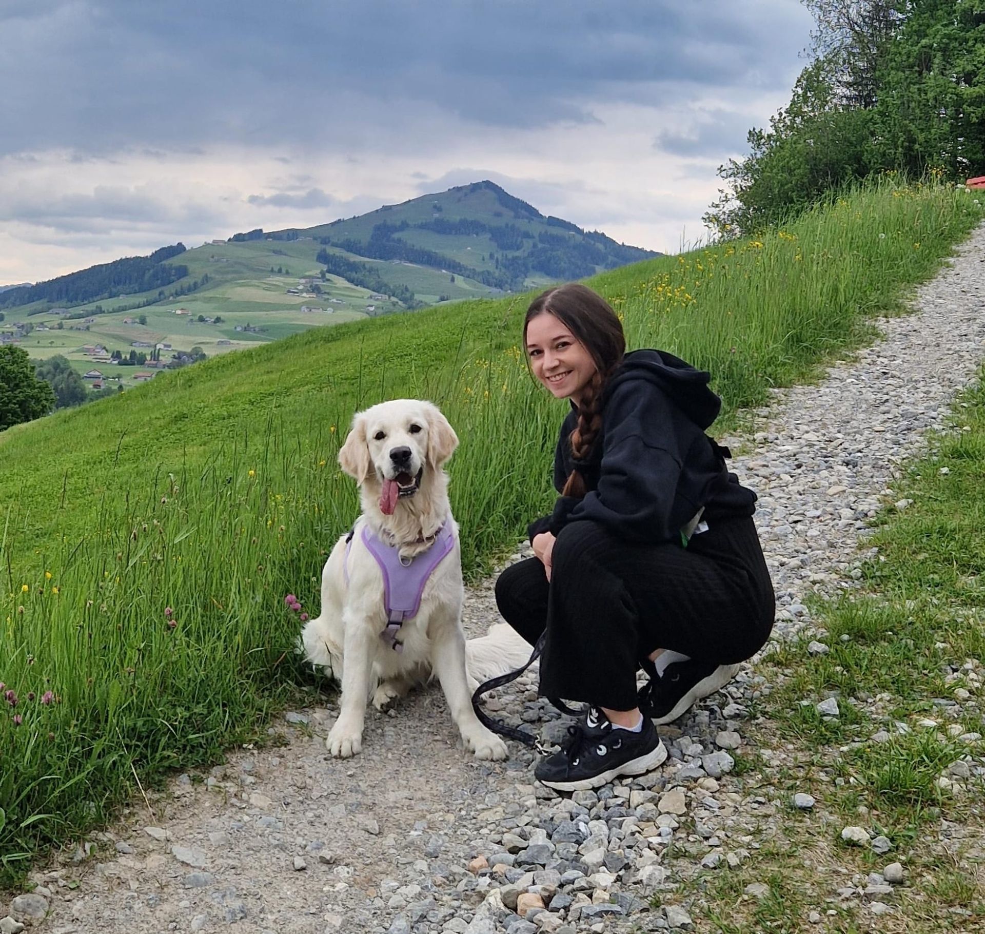 Frau hockt mit einem Golden Retriever auf einem Schotterweg, im Hintergrund grüne Hügel und Berge.
