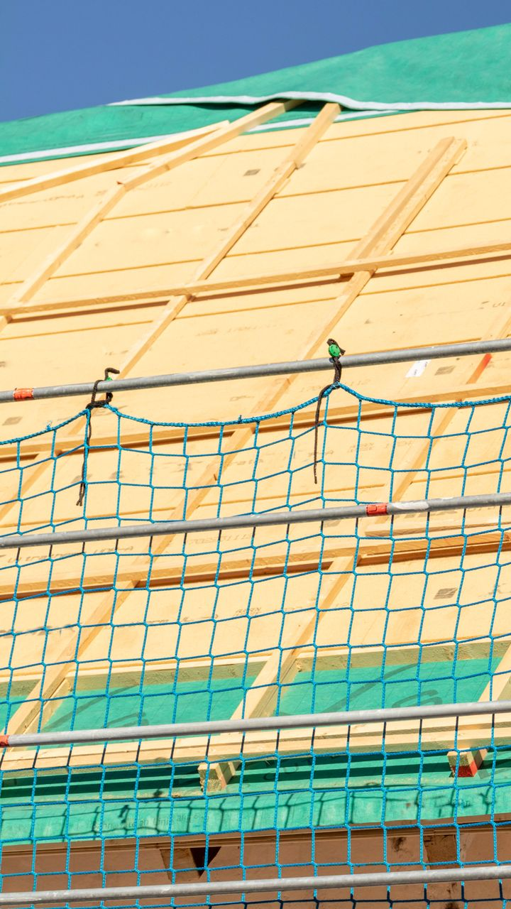 Chantier de construction avec filet de sécurité devant les chevrons du toit recouverts de contreplaqué et d'une bâche verte.