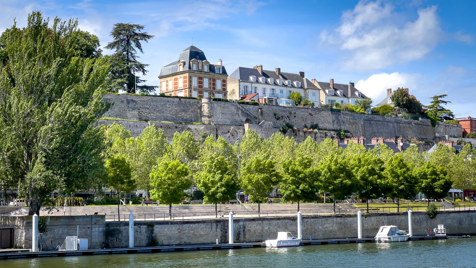 Cours d'eau en premier plan avec des bateaux amarrés le long d'un muret, en second plan se trouve de grosses maisons entourées d'arbres