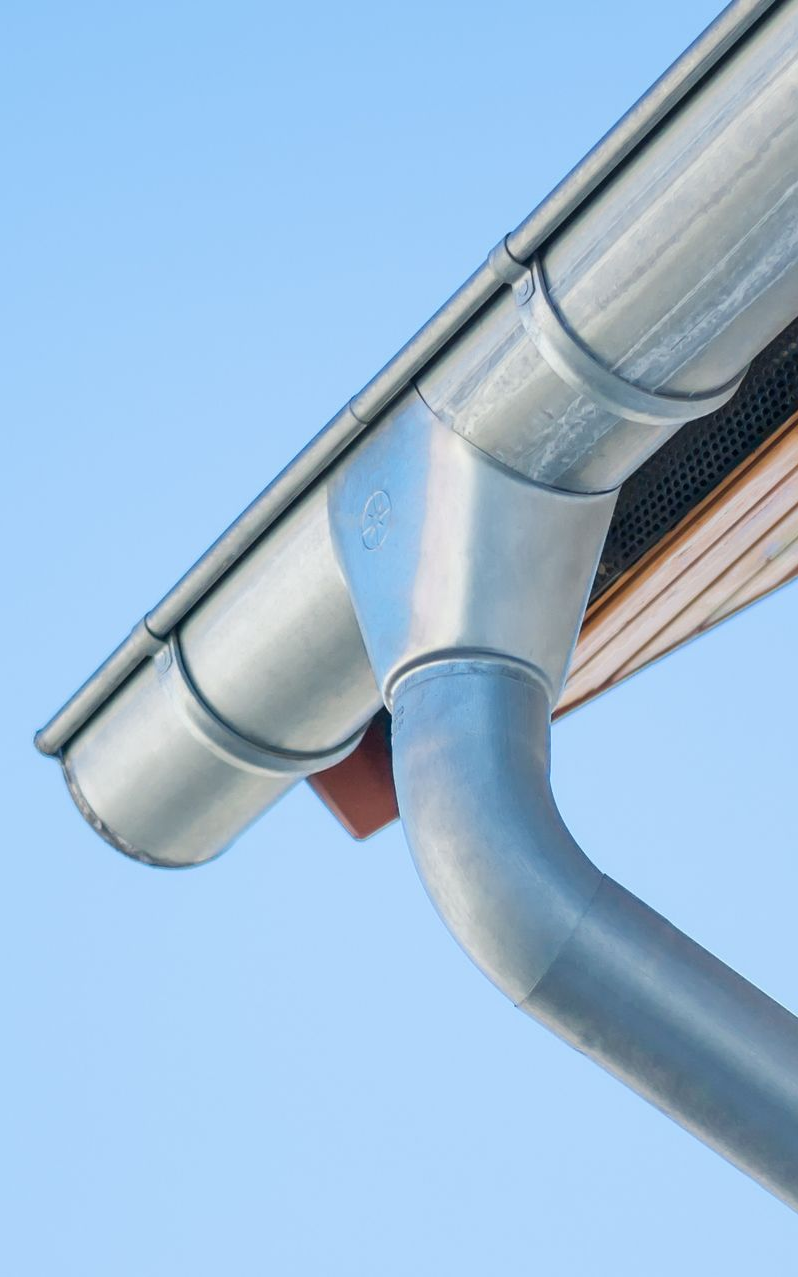 Gouttière et descente pluviale grises sur une maison blanche, se détachant sur un ciel bleu.