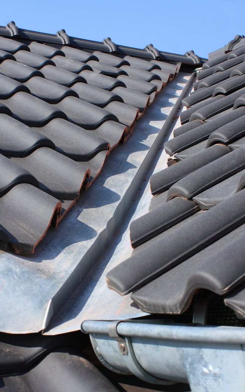 Des tuiles grises se chevauchent et rejoignent une noue métallique. Une gouttière est visible en bas.
