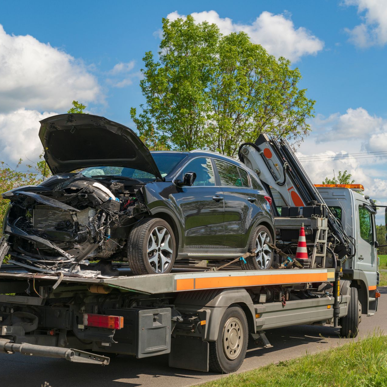 Une voiture noire gravement endommagée est remorquée. L'avant est écrasé, le capot est ouvert. En extérieur, par une journée ensoleillée.