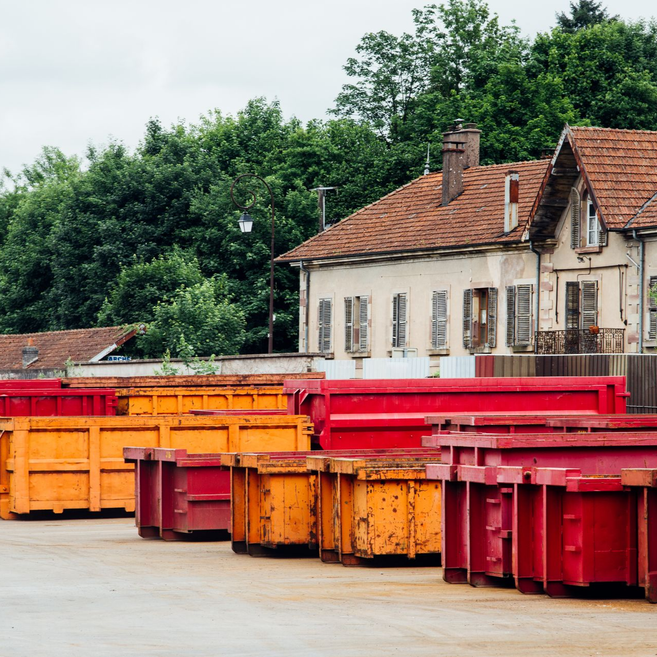 Des rangées de bennes à ordures orange et rouges devant un bâtiment délabré au toit de tuiles rouges, entouré d'arbres.