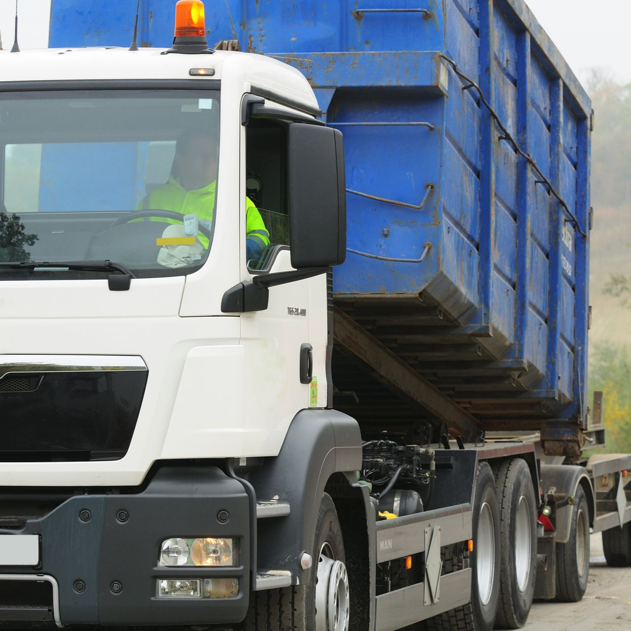 Camion blanc et bleu transportant un grand conteneur bleu, conducteur visible dans la cabine.