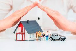 Unas manos forman un refugio protector sobre una casa en miniatura, una familia de figuritas y un coche de juguete sobre un fondo blanco.