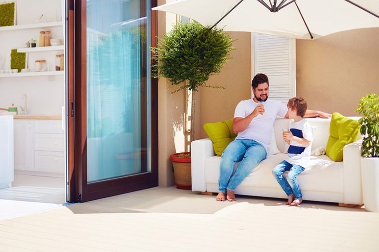 Un padre y su hijo están sentados en un sofá blanco de exterior en un patio, charlando y sosteniendo vasos bajo una sombrilla.