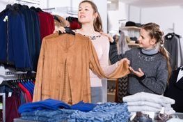 Una mujer sostiene una chaqueta color canela mientras compra con una niña en una tienda de ropa.