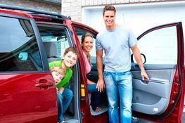 Una familia sonriente posa junto a un coche rojo con las puertas abiertas en la entrada de una casa.