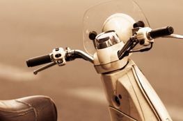 Primer plano, en tono sepia, del manillar, el velocímetro y el pequeño parabrisas de una scooter antigua.