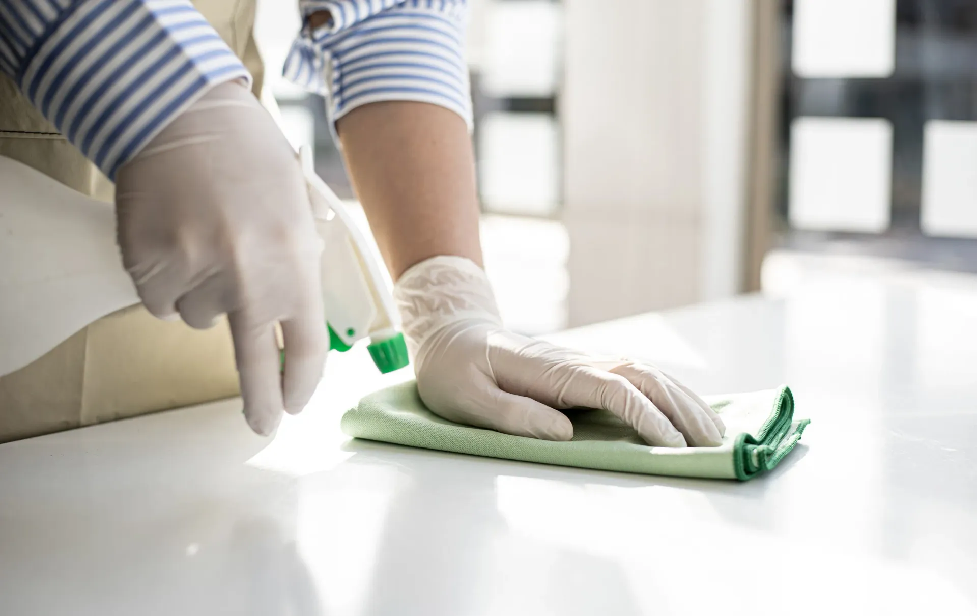 Persona con guantes, limpiando una superficie blanca con un paño verde y una botella de spray.