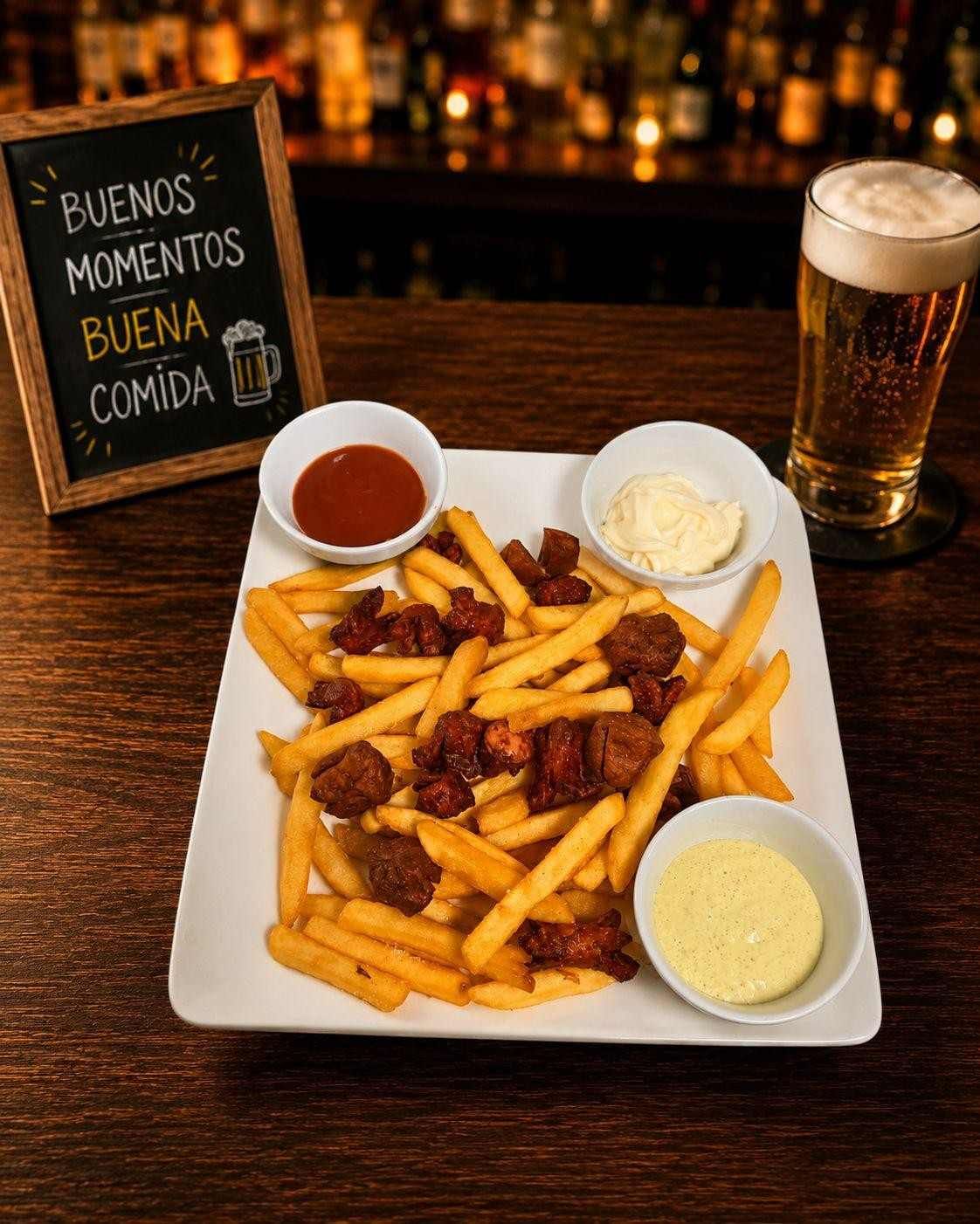 Plato de patatas fritas con tres salsas para mojar y una cerveza sobre una mesa de bar, con un pequeño cartel al fondo.