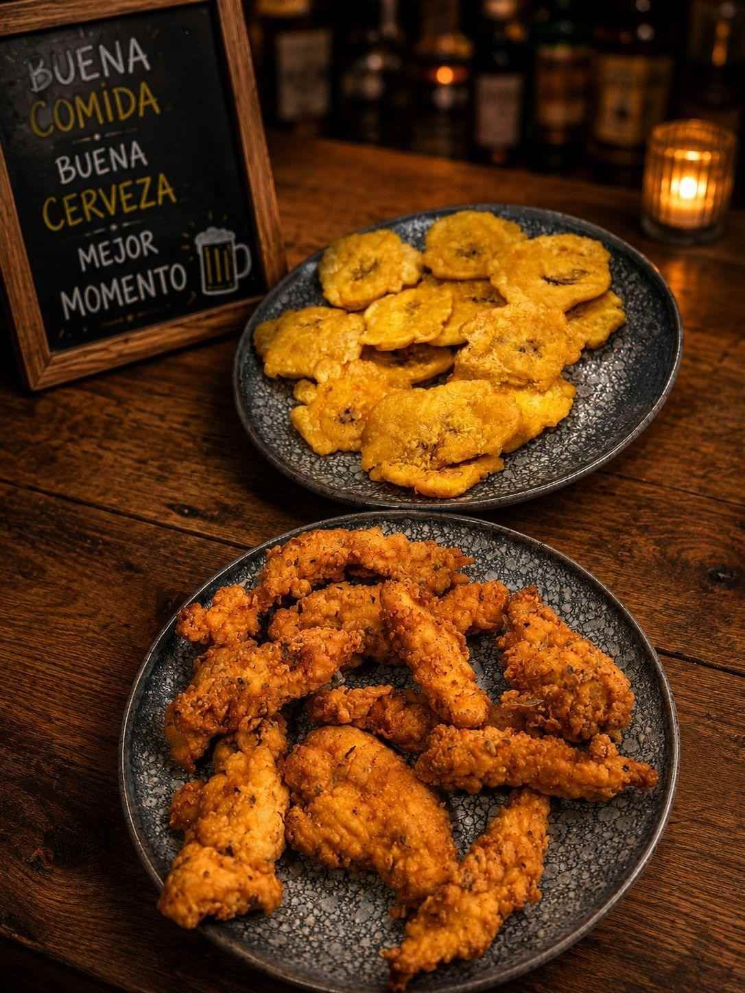 Two plates of seasoned fried snacks on a rustic table beside a sign reading “Buena comida, buena cerveza, mejor momento.”