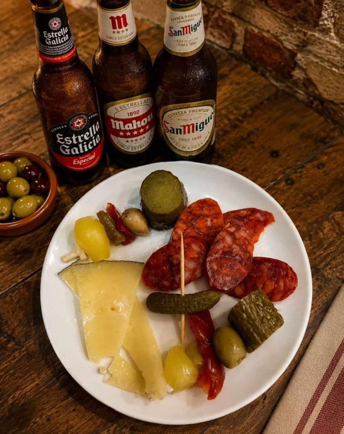 Plato con una selección de quesos, embutidos y encurtidos, acompañado de tres botellas de cerveza sobre una mesa de madera.