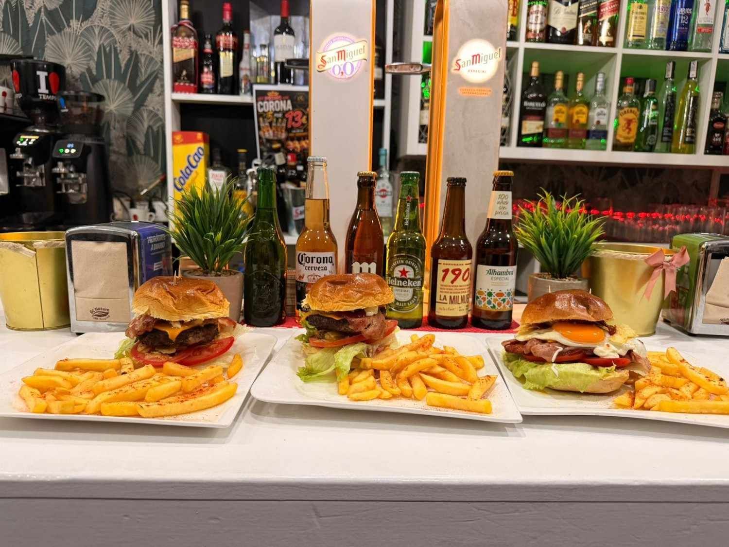 Tres hamburguesas con patatas fritas en platos blancos en la barra de un restaurante, con botellas y bebidas detrás.