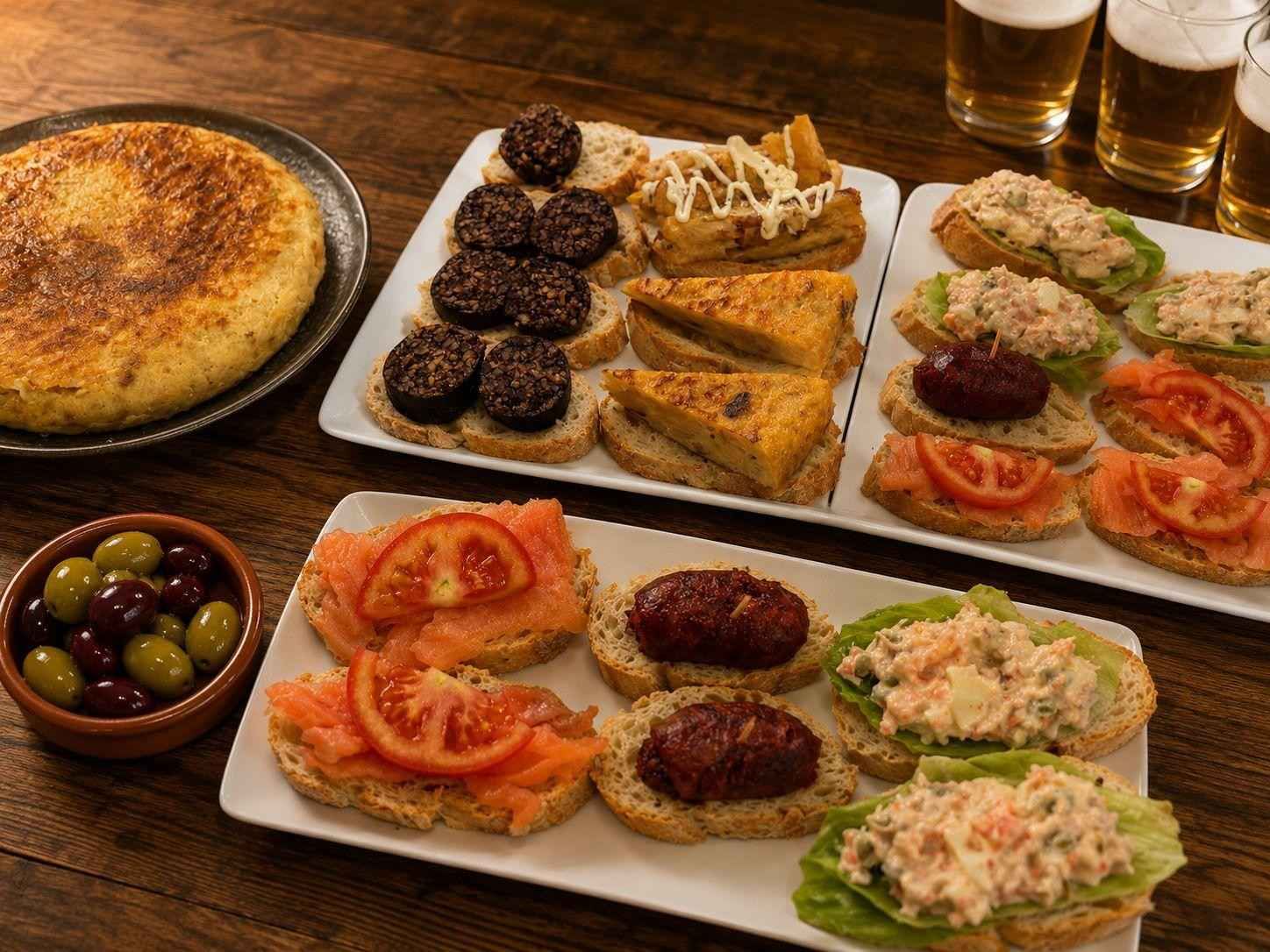 Surtido de aperitivos mediterráneos en bandejas, que incluyen salsa para untar, aceitunas, tomates rellenos y croquetas sobre una mesa de madera.
