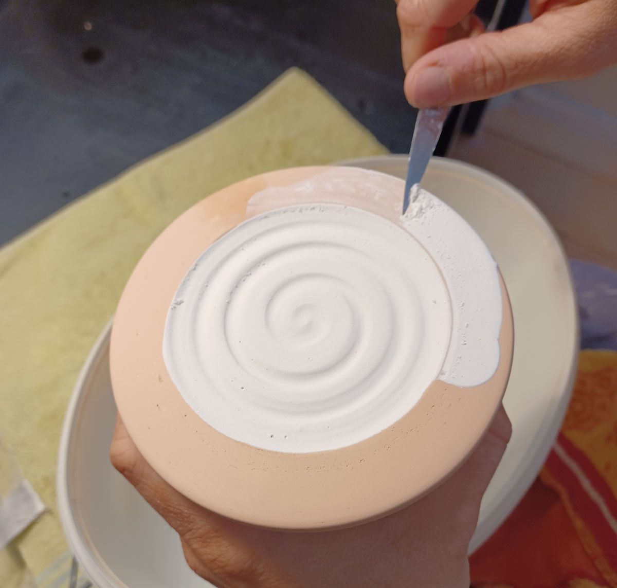 Hände schnitzen mit einem Werkzeug ein Spiralmuster in einen weißen Tontopf.