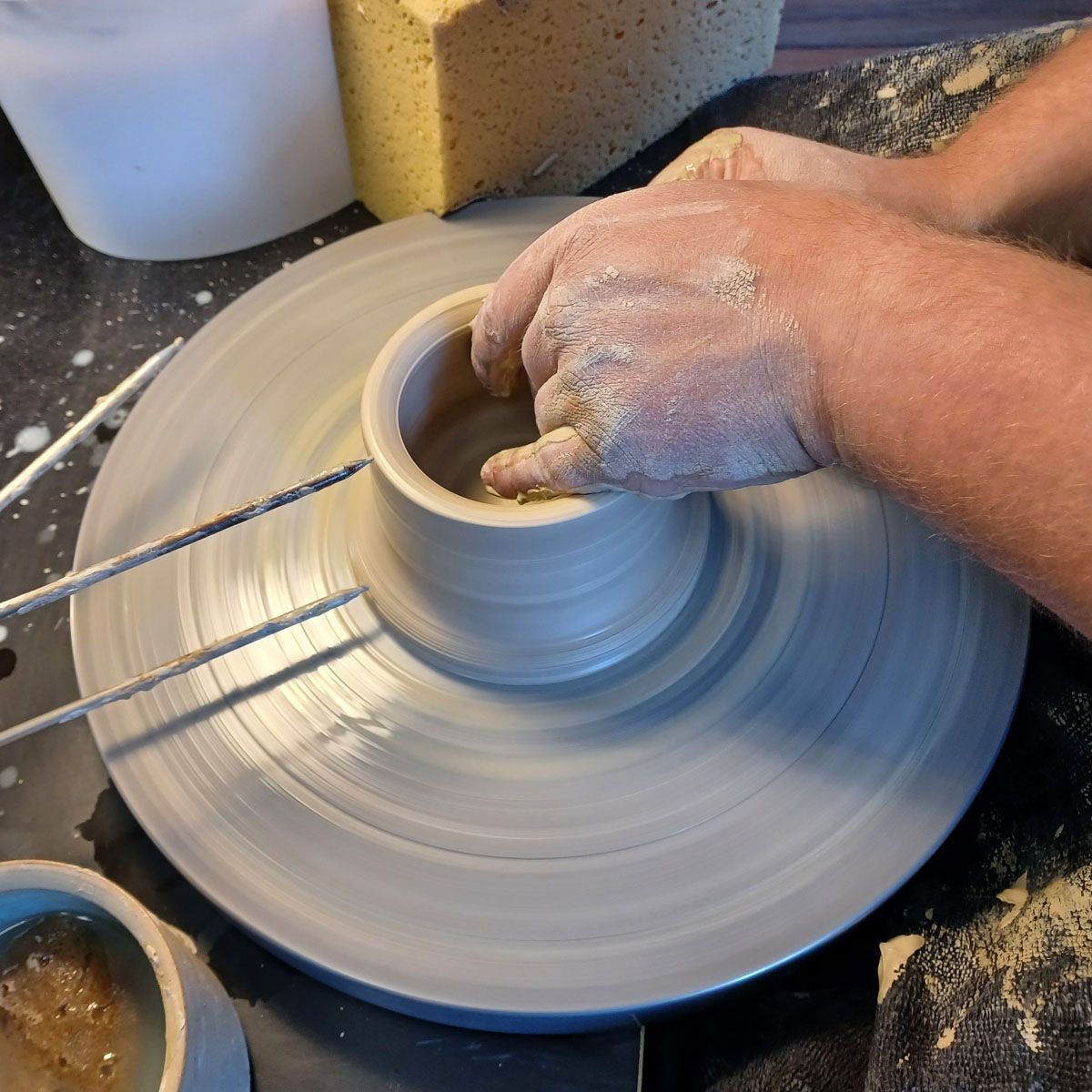 Hände formen Ton auf einer sich drehenden Töpferscheibe, umgeben von Spritzern nassen Tons in einem Atelier.