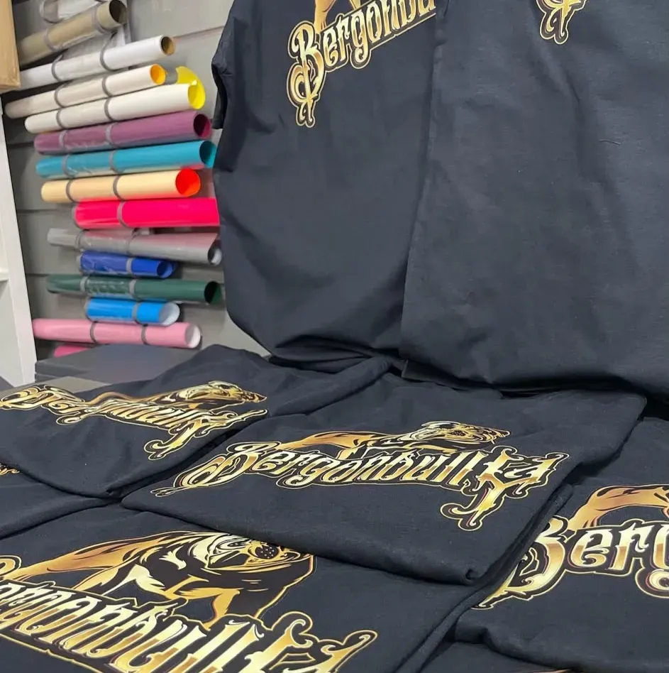 Camisetas negras apiladas con letras doradas ornamentadas junto a rollos de vinilo de colores en una imprenta.