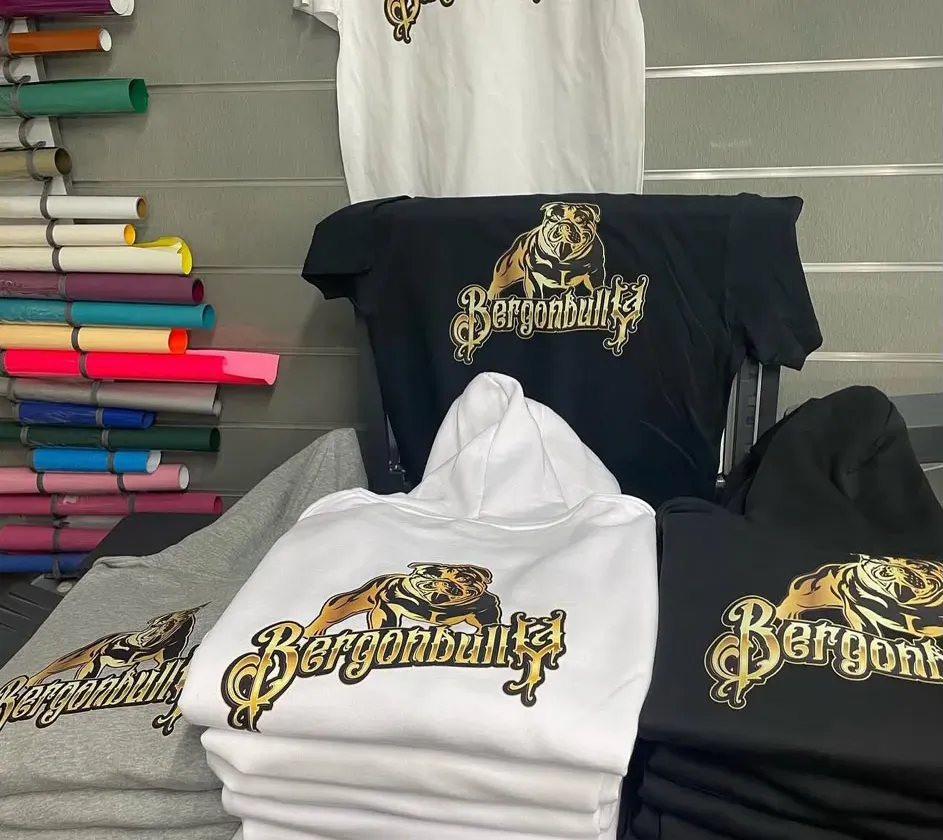 Sudaderas con capucha y camisetas estampadas en negro, blanco y gris con letras doradas, dobladas y colocadas en estantes de exhibición.