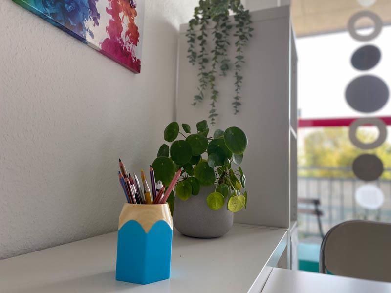 Ein bleistiftförmiger Behälter mit Bleistiften gefüllt steht auf einem Schreibtisch neben einer Topfpflanze