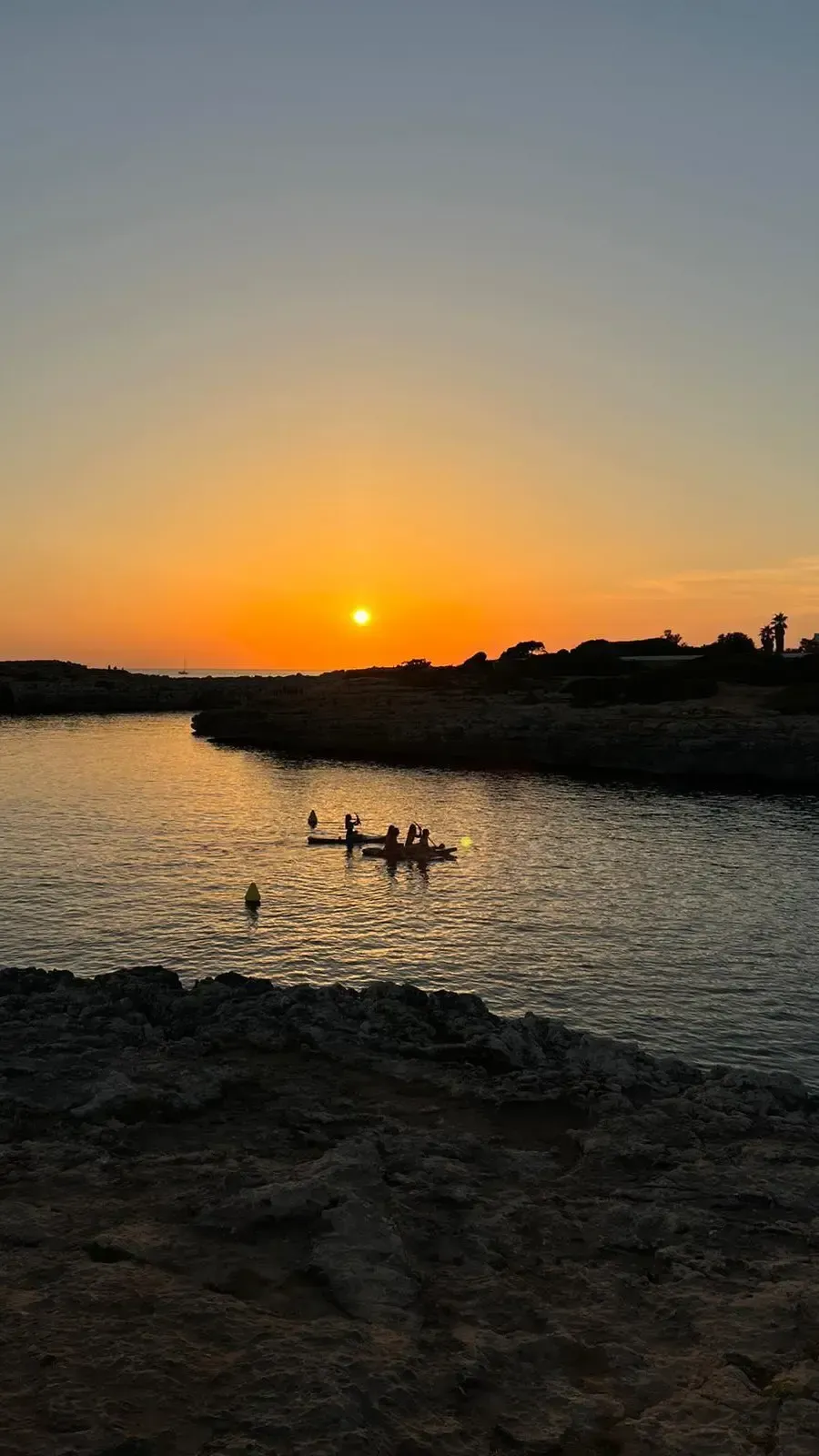 Gente practicando kayak en una ensenada tranquila durante una puesta de sol dorada, con la costa rocosa como telón de fondo.