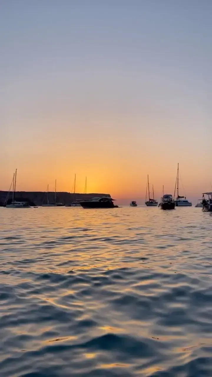 Barcos anclados en aguas tranquilas bajo una puesta de sol dorada, con un acantilado oscuro recortado contra el horizonte.
