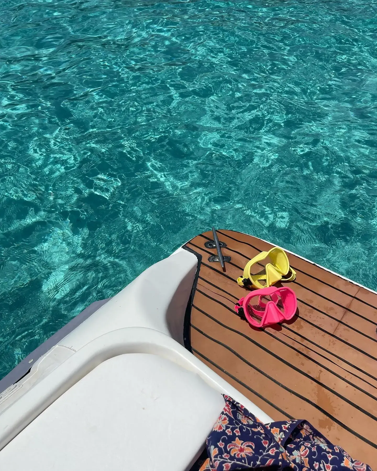 Máscaras de snorkel amarillas y rosas reposan sobre la cubierta de madera de un barco con vistas a aguas turquesas brillantes y cristalinas.