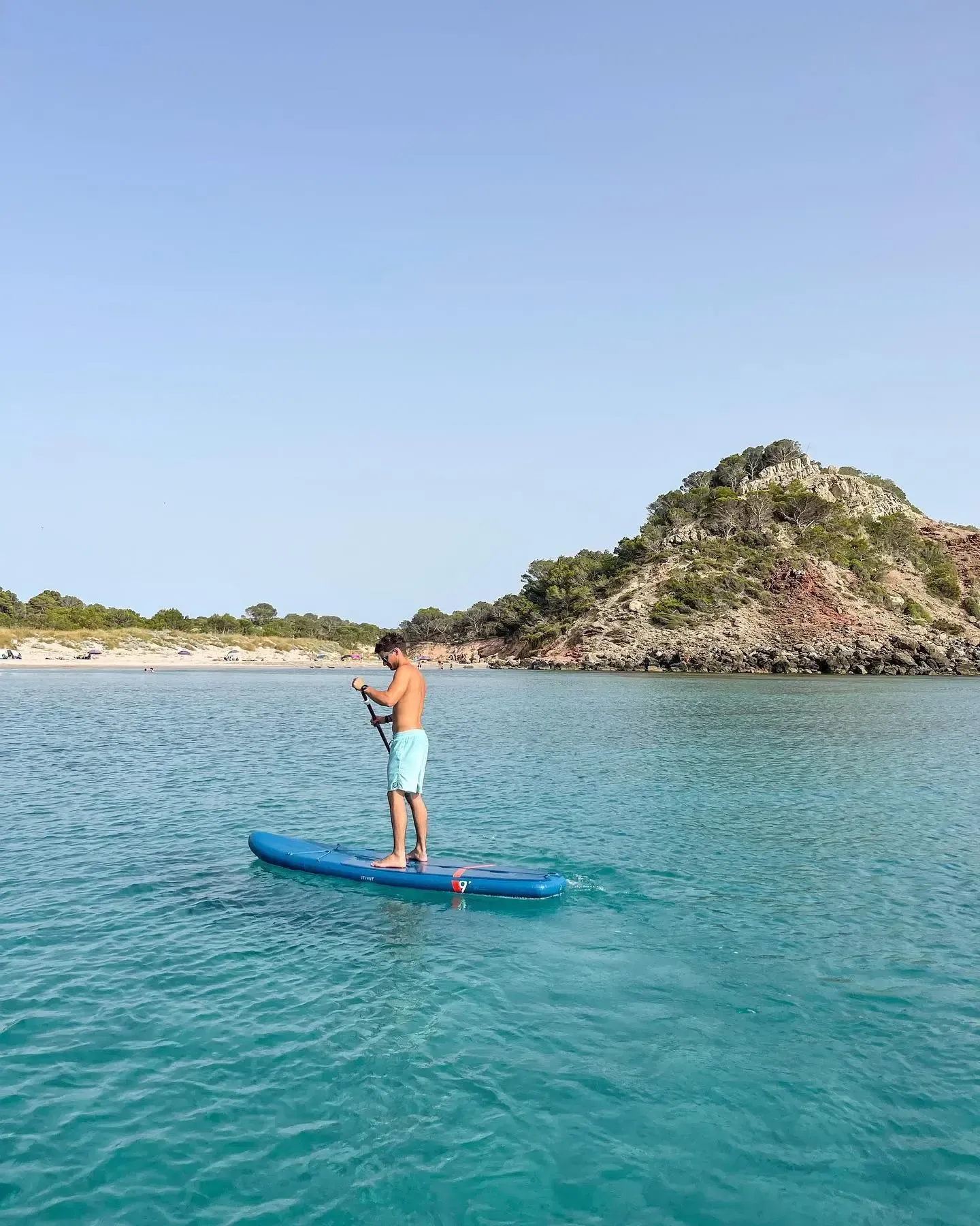 Una persona de pie sobre una tabla de paddle surf azul en aguas cristalinas de color turquesa, con una colina rocosa cubierta de árboles al fondo.