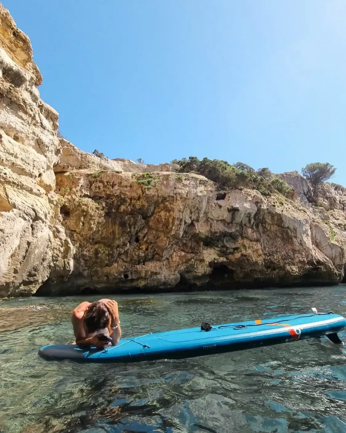 Una persona yace sobre una tabla de remo azul en aguas claras y poco profundas, al pie de un acantilado rocoso, bajo un cielo azul brillante.